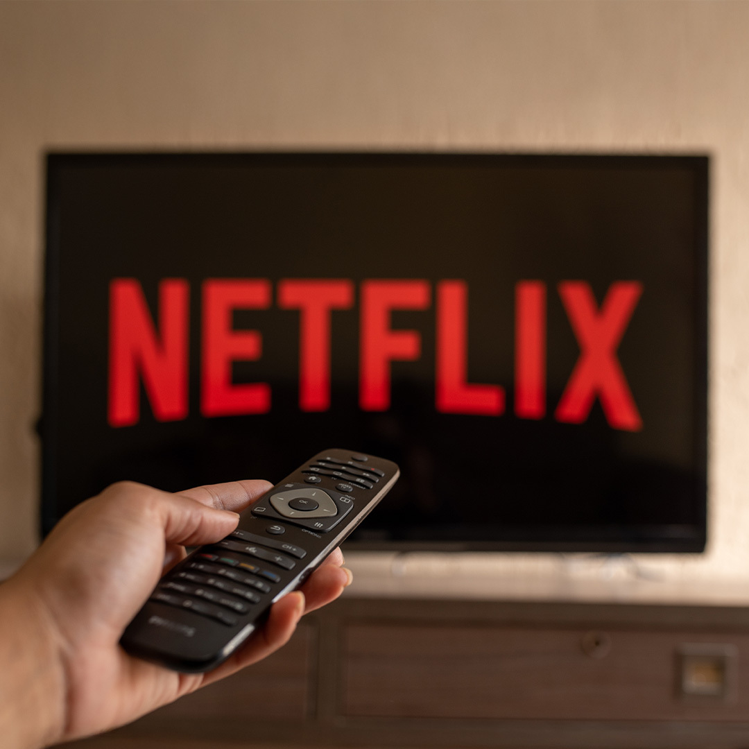 Logo Netflix à l'écran d'un téléviseur en arrière-plan et une main tenant une télécommande en avant-plan.