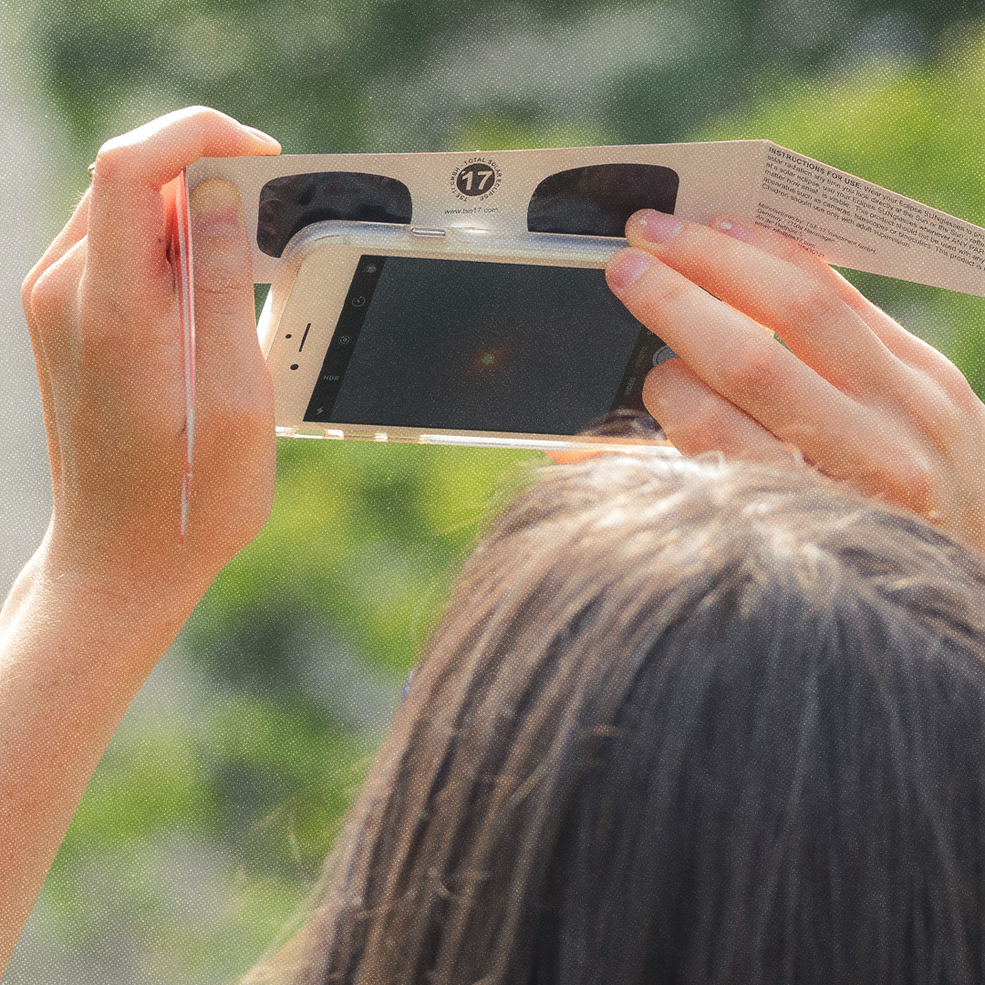 Une femme prend une photo d'une éclipse solaire en utilisant son cellulaire et une paire de lunettes pour éclipse solaire.