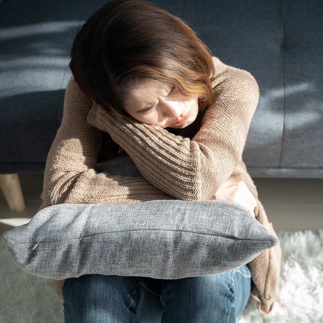 A woman sits on the floor by the couch looking sad.