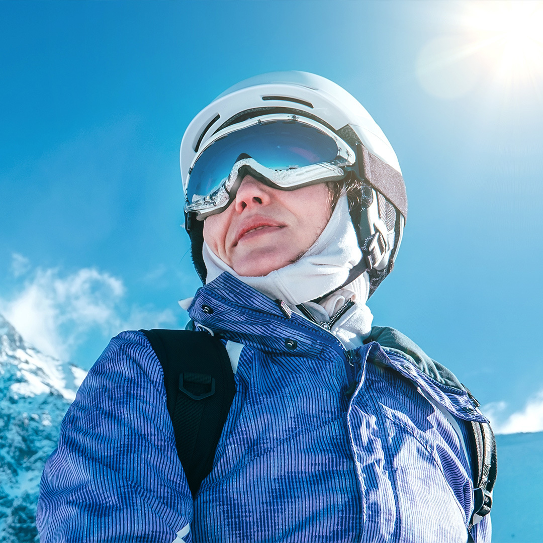 A skier wearing a helmet and goggles stands in front of a backdrop of snowy mountains.