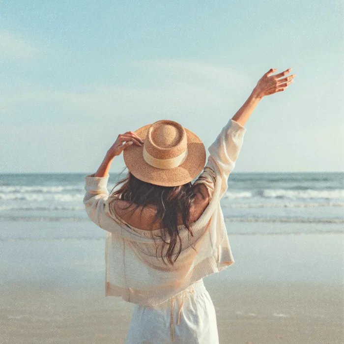 Une femme de dos portant un chapeau, les bras levés, face à la mer.